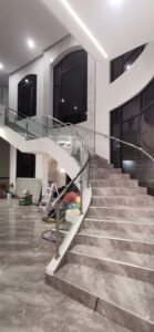 Interior lobby with a curved marble staircase and glass railing; construction workers on the floor nearby with tools and materials tall windows in the background