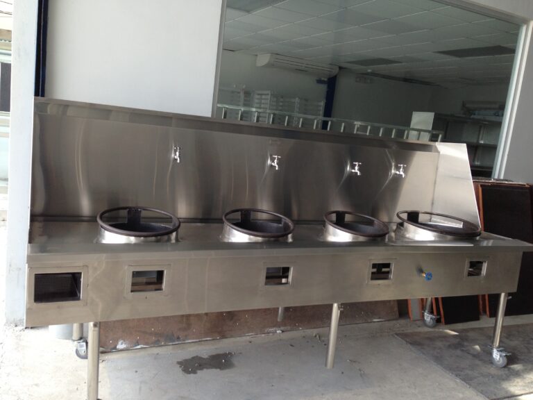 Industrial stainless-steel commercial kitchen unit with four circular open burner rings and faucet controls, on a wheeled base in a kitchen area