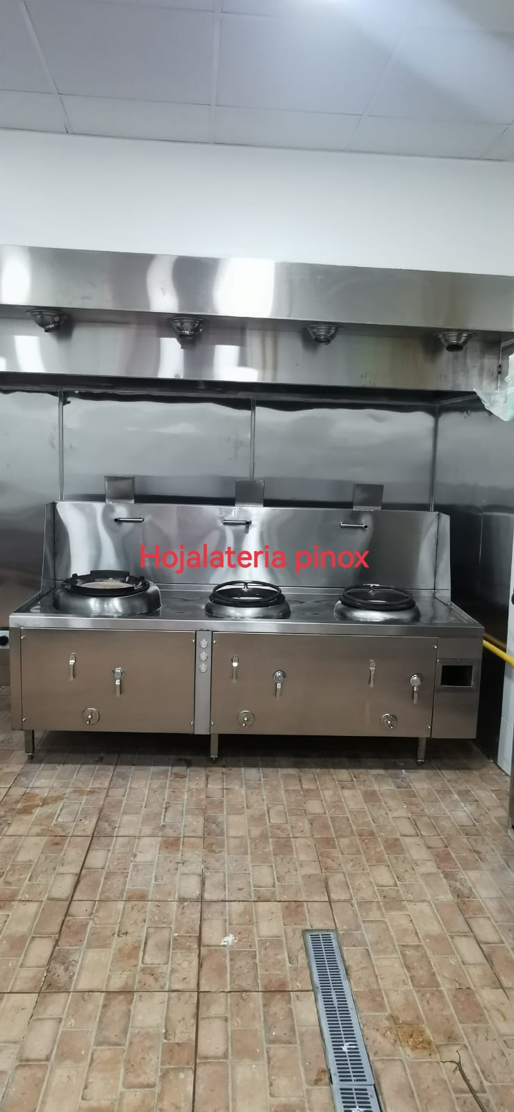 Industrial stainless-steel three-burner stove with cabinet bases in a commercial kitchen setting, brand text 'Hojalateria pinox' visible on the backsplash