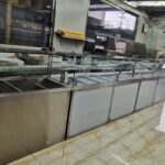 Empty stainless steel refrigerated display counter with glass sneeze guard inside a storefront deli/kitchen area windowed to street outside
