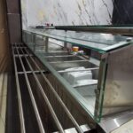 Stainless steel and glass food display counter with a marble-patterned wall behind it in a modern shop setting.
