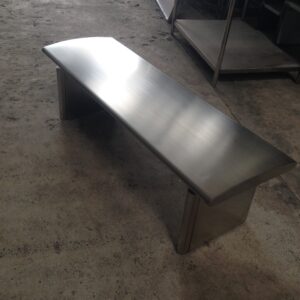 Brushed stainless steel bench in an industrial workshop on a concrete floor. A metal cabinet/part of a stainless steel shelving unit is visible in the background.