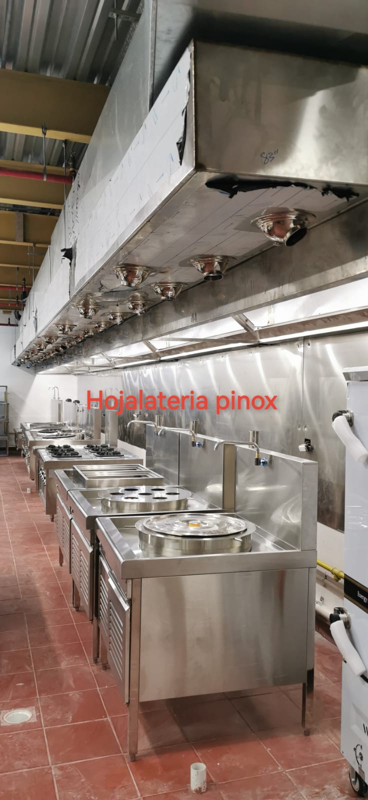 Commercial stainless steel kitchen with a long row of stoves and a large vent hood; red text 'Hojalateria pinox' on the back wall.