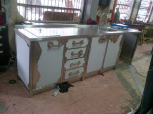 Industrial kitchen counter with stainless steel top, damaged cabinet doors, and scattered debris on a tiled floor; cluttered workspace.