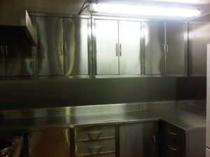 Row of tall stainless steel cabinets with curved handles along a commercial kitchen wall under bright lighting.