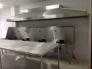 Industrial kitchen with large stainless steel vent hood and grilling/food prep station covered in plastic wrap.