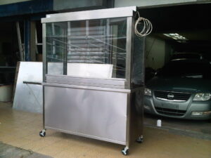 Stainless steel mobile display cabinet with glass front and shelves in a dim garage, with a coiled cable hanging on the right.