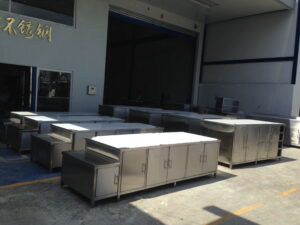 Row of stainless steel kitchen cabinets and worktables arranged outdoors beside an industrial building.