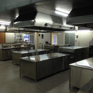 Industrial commercial kitchen with stainless steel worktables, cabinets, and large overhead ventilation ducts under a dark ceiling lighting fixtures.